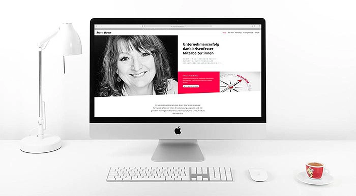 Eleganter weißer Schreibtisch mit einem iMac, der eine Website mit einem Schwarz-Weiß-Porträt anzeigt; links eine Lampe, rechts eine Kaffeetasse.