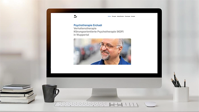 Eleganter weißer Schreibtisch mit einem iMac, der eine Website mit einem Porträt anzeigt; links ein Bücherstapel und eine schwarze Tasse, rechts eine Stiftebox.